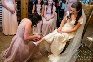 A bride sitting in a chair has her bridesmaid slips her shoes on her feet.