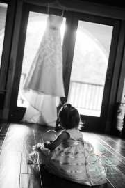 A tiny little flower girl playing on the floor in front of the wedding dress which is hanging overhead on the door.