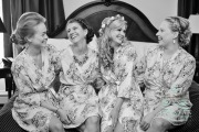A bride laughing with her bridesmaids as they sit in their floral bridal robes before the wedding.