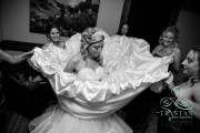 A photograph of a bride looking like a white flower with the dress spread out around her like petals as the bridesmaids try to fasten the ribbons and corset underneath before the wedding.