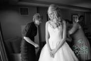 A bride stands patiently waiting with her hands folded as her mother inspects the wedding dress before the wedding.