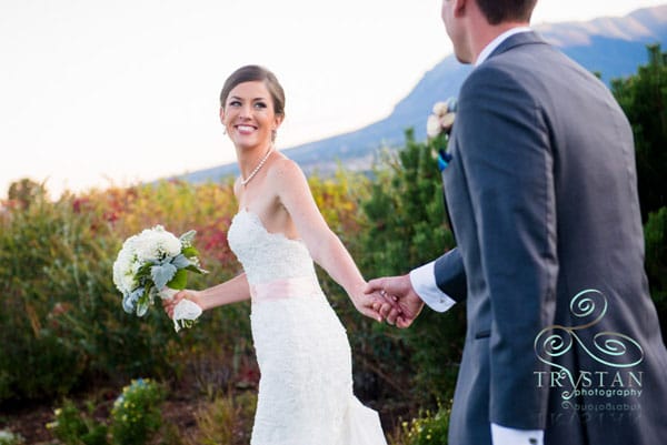 A photograph of a bride looking back over her shoulder and smiling at the groom as they walk away while holding hands.