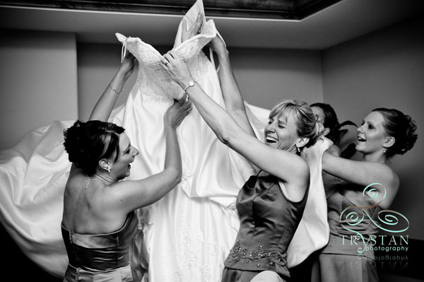 A photograph of a bride's mom and her bridesmaids laughing as they try to get her into the wedding dress.