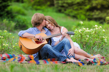 A portrait of a couple cuddling together in a lush green field, she is smiling and resting her head on his shoulder while he is playing guitar.