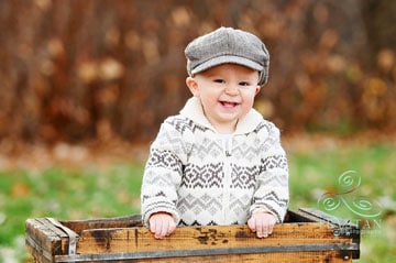 A portrait of a little boy in an adorable paper-boy hat and sweater laughing in an antique crate in a park in Autumn in Colorado Springs..