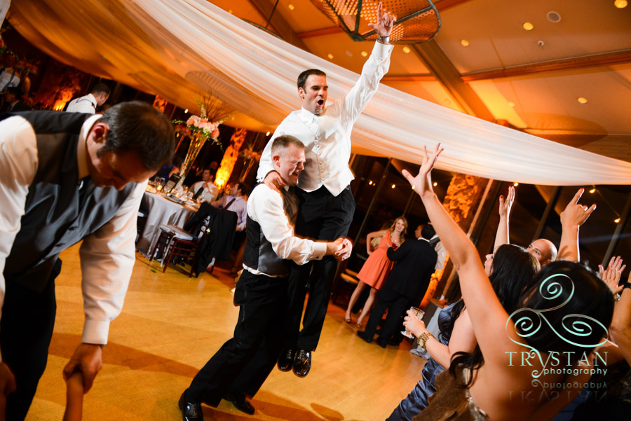 A photograph of a groomsman lifting the groom in the air at the reception and the groom has his hand up and is smiling and yelling in joy.