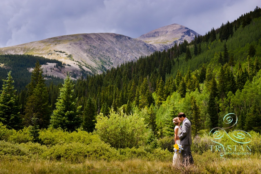 A wedding photograph of a bride and groom kissing in a valley meadow with a mountain peak rising up behind them near Breckenridge, Colorado.