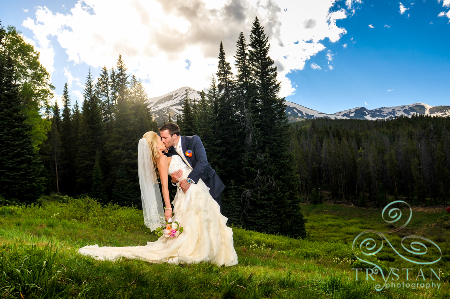 A photograph of a groom kissing and dipping his bride dramatically on a mountainside with Peak 9 looming behind them.